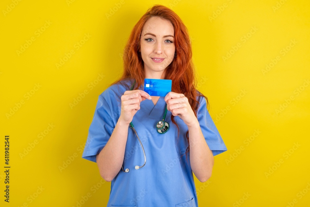 Photo of young red-haired doctor woman over yellow studio background positive smile hold credit card income salary Photo of young red-haired doctor woman over yellow studio background positive smile hold credit card income salary