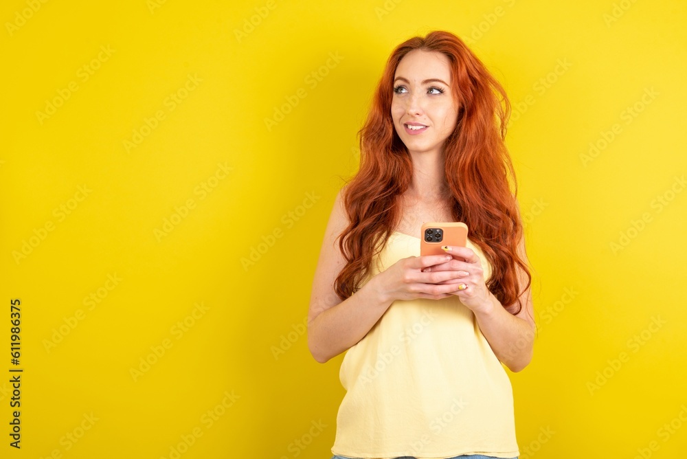 young beautiful red haired woman wearing yellow blouse over yellow studio background holding in hands mobile phone and looking aside with dreamy look. young beautiful red haired woman wearing yellow blouse over yellow studio background holding in hands mobile phone and looking aside with dreamy look.