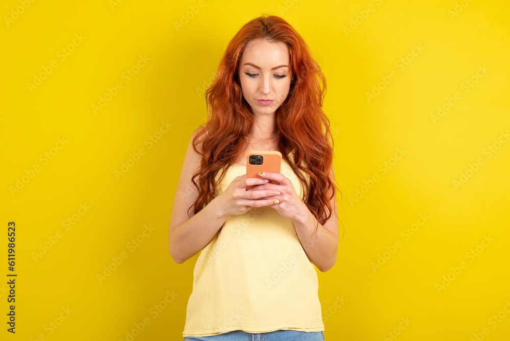 Focused young beautiful red haired woman wearing yellow blouse over yellow studio background use smartphone reading social media news, or important e-mail Focused young beautiful red haired woman wearing yellow blouse over yellow studio background use smartphone reading social media news, or important e-mail