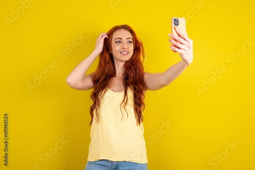 young beautiful red haired woman wearing yellow blouse over yellow studio background smiling and taking a selfie ready to post it on her social media.