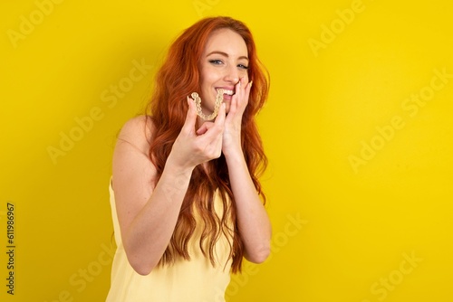 Happy young beautiful red haired woman wearing yellow blouse over yellow studio background holding and showing at camera an invisible aligner while laughing. Dental healthcare and confidence concept.