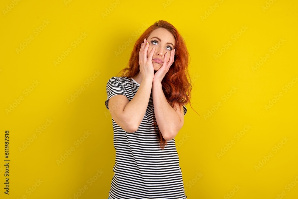 young beautiful red haired woman wearing striped t shirt over yellow studio background keeps hands on cheeks has bored displeased expression. Stressed hopeless model young beautiful red haired woman wearing striped t shirt over yellow studio background keeps hands on cheeks has bored displeased expression. Stressed hopeless model
