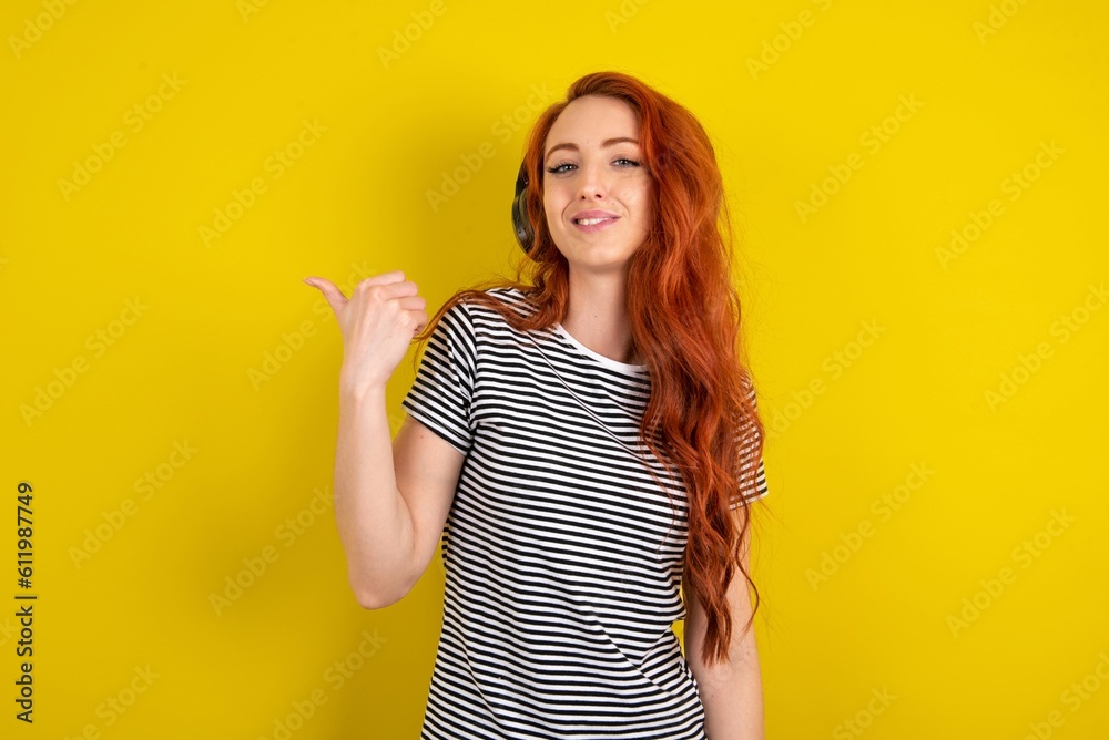 young beautiful red haired woman wearing striped shirt over yellow studio background listens audio track via wireless headphones points thumb away advertises copy space young beautiful red haired woman wearing striped shirt over yellow studio background listens audio track via wireless headphones points thumb away advertises copy space