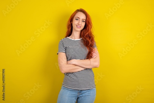 Portrait of charming young beautiful red haired woman wearing striped shirt over yellow studio background standing confidently smiling toothily with hands folded