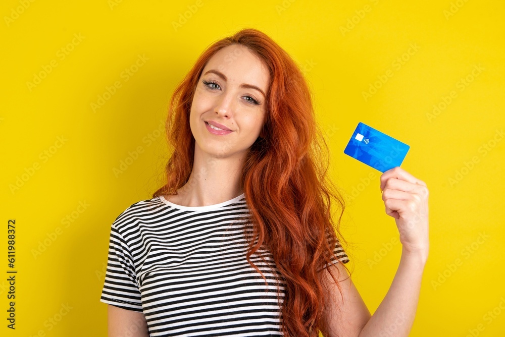 Close up photo of optimistic young beautiful red haired woman wearing striped shirt over yellow studio background hold card Close up photo of optimistic young beautiful red haired woman wearing striped shirt over yellow studio background hold card