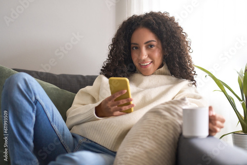 Young pretty latin woman sitting on couch using cell phone, holding smartphone looking at camera wit cellphone doing online shopping in mobile digital shop app, checking social media commercial offer.