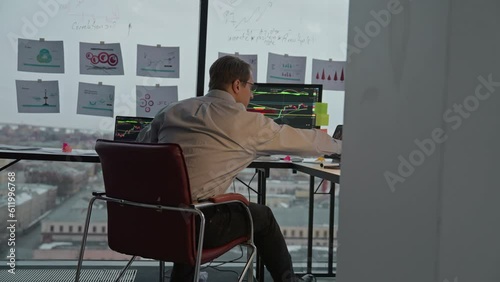 Businessman checks status of financial investments on computer and tablet in office at desk by panoramic window photo
