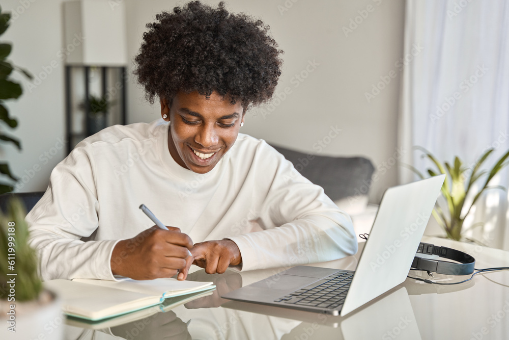 Happy African American teen student elearning at home on pc, writing notes. Smiling teenage boy using laptop watching webinar, hybrid learning english online virtual class, sitting at home table. Happy African American teen student elearning at home on pc, writing notes. Smiling teenage boy using laptop watching webinar, hybrid learning english online virtual class, sitting at home table.