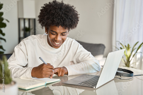 Happy African American teen student elearning at home on pc, writing notes. Smiling teenage boy using laptop watching webinar, hybrid learning english online virtual class, sitting at home table.