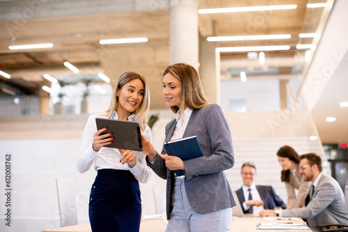 Two young business women looking at financial results on digital tablet in front of their team at the office