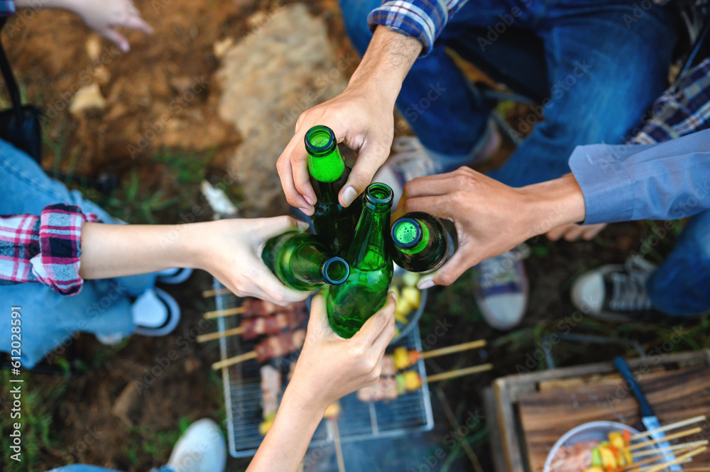 Top view shot of friends clinking bottle of beer during camping outdoor with barbecue on grill below in background. Celebrating summer camping party natural outdoor. Top view shot of friends clinking bottle of beer during camping outdoor with barbecue on grill below in background. Celebrating summer camping party natural outdoor.