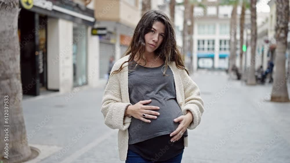 Young pregnant woman standing with serious expression touching belly at street Young pregnant woman standing with serious expression touching belly at street