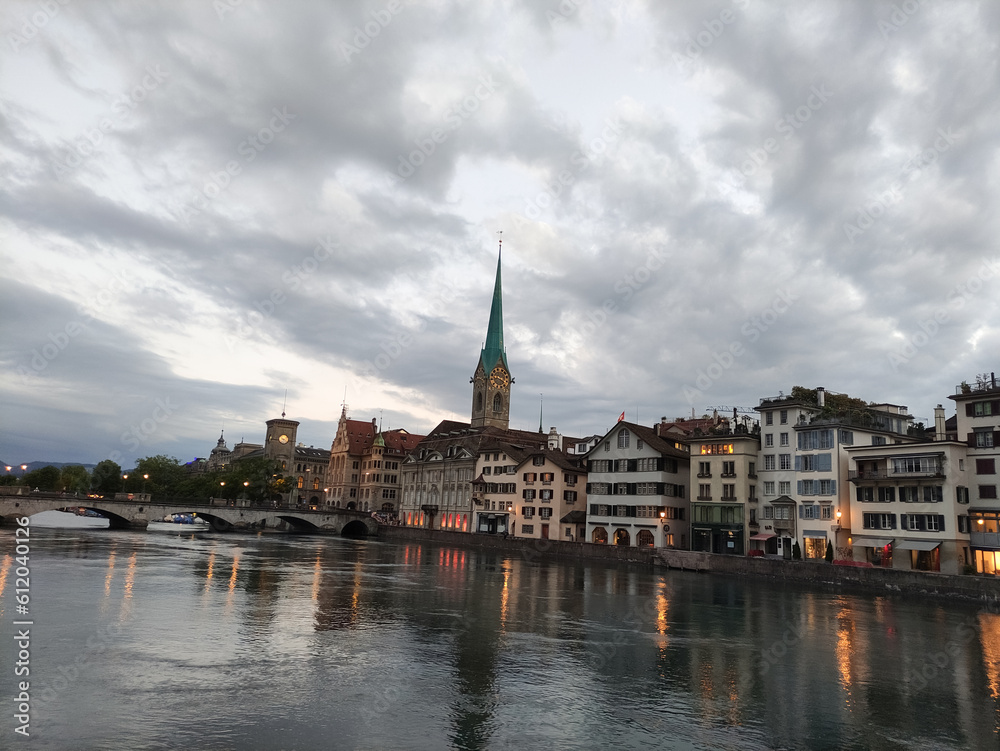 Zurich, Switzerland. River Limmat. Sunset sky, dark clouds, evening time. Orange lights, sundown. Old city centre, clock tower, urban landscape.