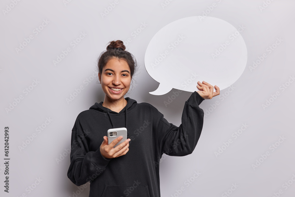 Smiling Latin woman holds aspeech bubble in one hand symbolizing her thoughts and ideas with other hand gripping smartphone represents perfect balance between introspection and connectivity. Smiling Latin woman holds aspeech bubble in one hand symbolizing her thoughts and ideas with other hand gripping smartphone represents perfect balance between introspection and connectivity.