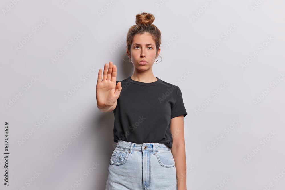 Young woman exudes seriousness as she confidently displays stop gesture with her palm conveying strong message of authority and determination asks to halt dressed in black t shirt and jeans.