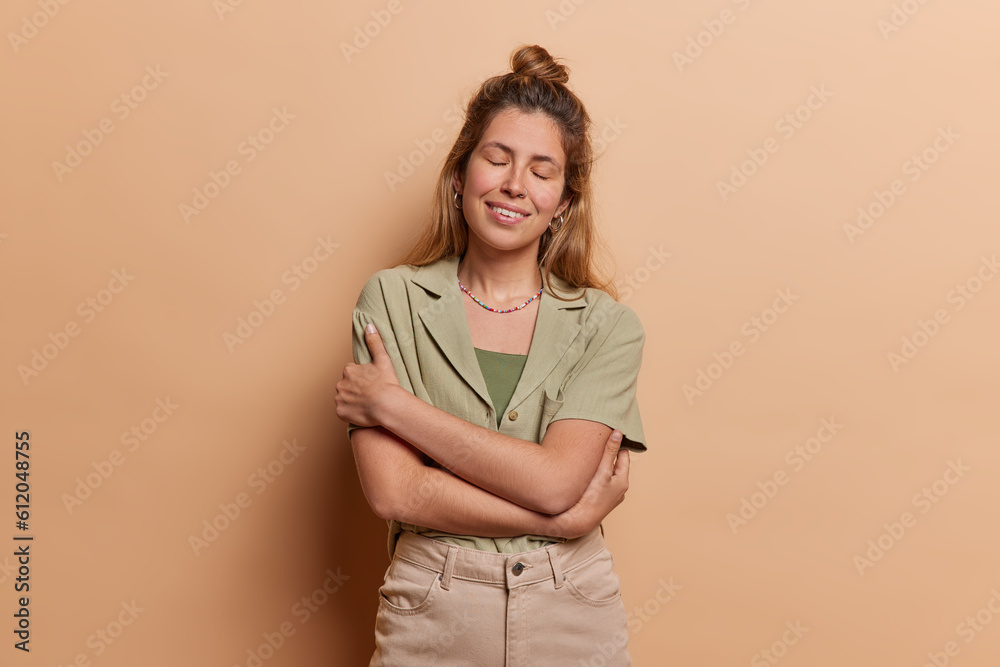 Obraz premium European girl wears shirt trousers and embraces herself with warm smile sstands isolated against beige background emanating sense of inner peace and self acceptance isolated over brown background