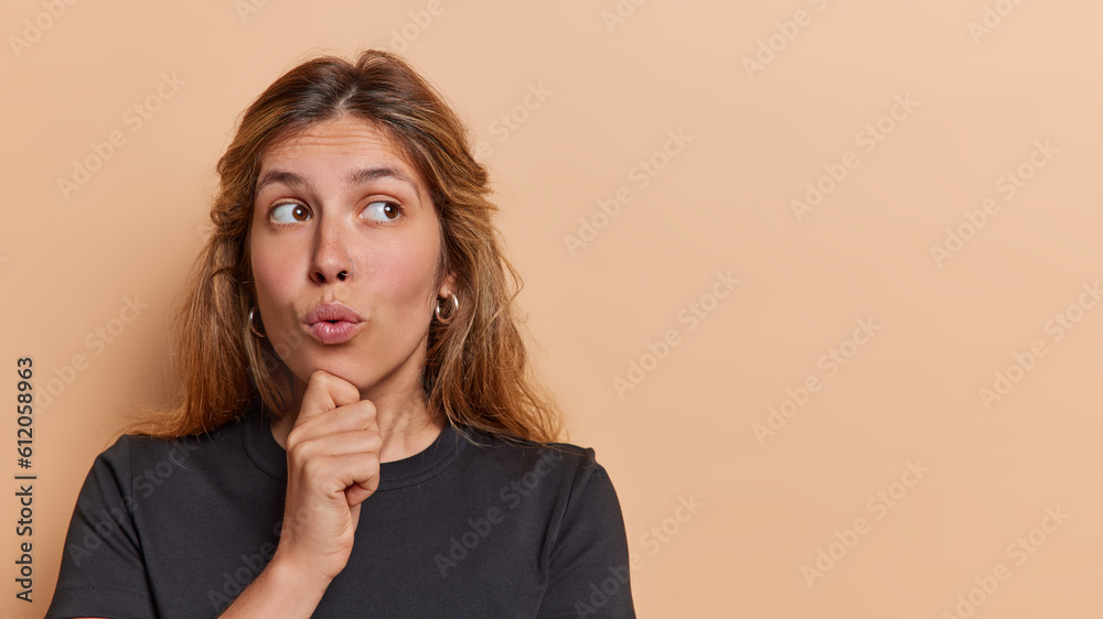 Horizontal shot of European woman keeps hand on chin concentrated aside has widely opened eyes dressed in black t shirt isolated over brown background copy space for your advertising content Horizontal shot of European woman keeps hand on chin concentrated aside has widely opened eyes dressed in black t shirt isolated over brown background copy space for your advertising content