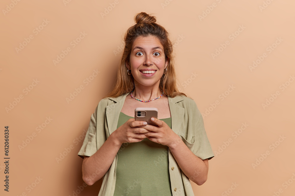 Beautiful female model is struck with amazement as she receives text message on her mobile phone smiles broadly dressed in shirt isolated over brown background. People technology and communication Beautiful female model is struck with amazement as she receives text message on her mobile phone smiles broadly dressed in shirt isolated over brown background. People technology and communication