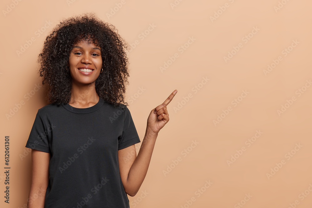 Young African woman stands against brown backdrop pointing her fingers away to draw attention to compelling copy space has engaging expression and confident posture wears casual black t shirt Young African woman stands against brown backdrop pointing her fingers away to draw attention to compelling copy space has engaging expression and confident posture wears casual black t shirt