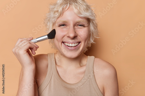 Horizontal shot of fair haired woman radiates joy looks directly at camera holds makeup brush in hand applies powder on cheek smiles broadly shows white teeth wears tshirt isolated on brown wall