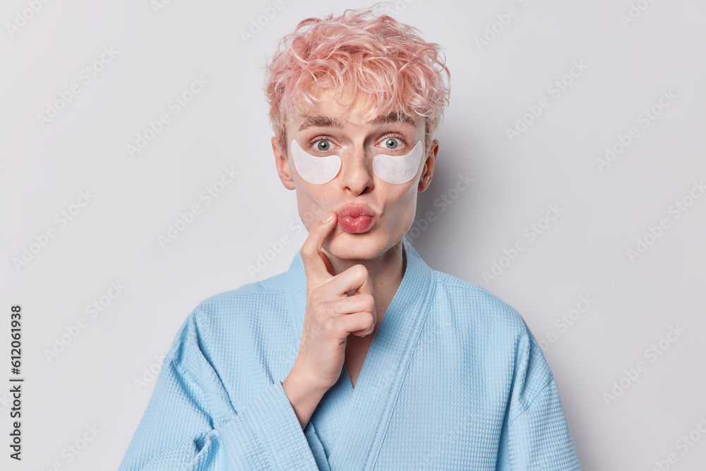 Surprised pink haired man gently applies cosmetic patches follows his morning routine has lips rounded keeps finger near mouth dressed in comfortable blue robe isolated over white background.
