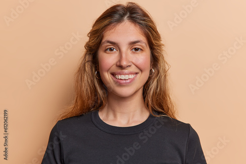 Portrait of lovely long haired woman smiles broadly shows white teeth has piercing in nose dressed in casual black t shirt isolated over brown background expresses positive emotions. Happiness concept