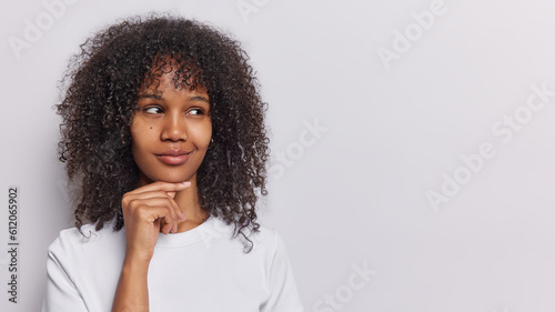 Studio shot of pretty woman with dark curly hair keeps hand under chin daydreams about something thinks about pleasant has tricky plan dressed in casual t shirt isolated on white background copy space
