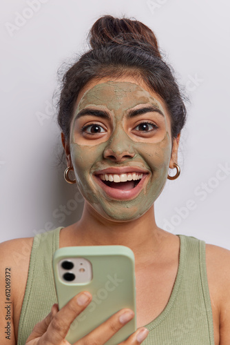 Brunette woman with hair bun smiles broadly as she checks and receives message on her mobile phone applies nourishing beauty mask on face aims to improve skin condition wears t shirt isolated on white photo