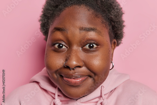 Close up shot of dark skinned chubby woman smiles pleasantly has piercing in nose dressed in casual hoodie hears something with curious expression isolated over pink background. Human reactions