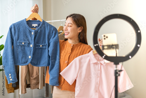 Vlogger, blogger small SME business asian beautiful woman using mobile phone video call for sell clothes live stream selling online, show product present detail at home office, entrepreneur ecommerce