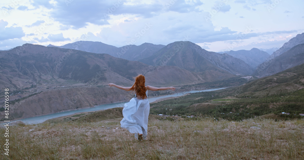 Young beautiful girl with red hair wearing white dress walking on top of a mountain facing wind blowing her hair and dress - freedom, adventure, harmony Young beautiful girl with red hair wearing white dress walking on top of a mountain facing wind blowing her hair and dress - freedom, adventure, harmony