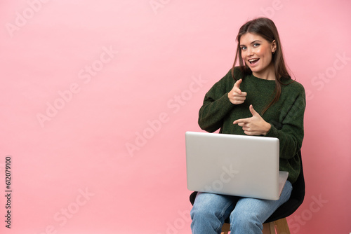 Young caucasian woman sitting on a chair with her laptop isolated on pink background pointing to the front and smiling