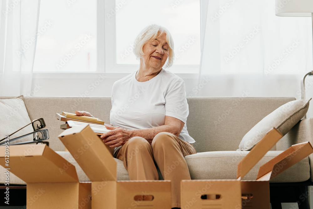 elderly woman sits on a sofa at home with boxes. collecting things with memories albums with photos and photo frames moving to a new place cleaning things and a happy smile. Lifestyle retirement. elderly woman sits on a sofa at home with boxes. collecting things with memories albums with photos and photo frames moving to a new place cleaning things and a happy smile. Lifestyle retirement.