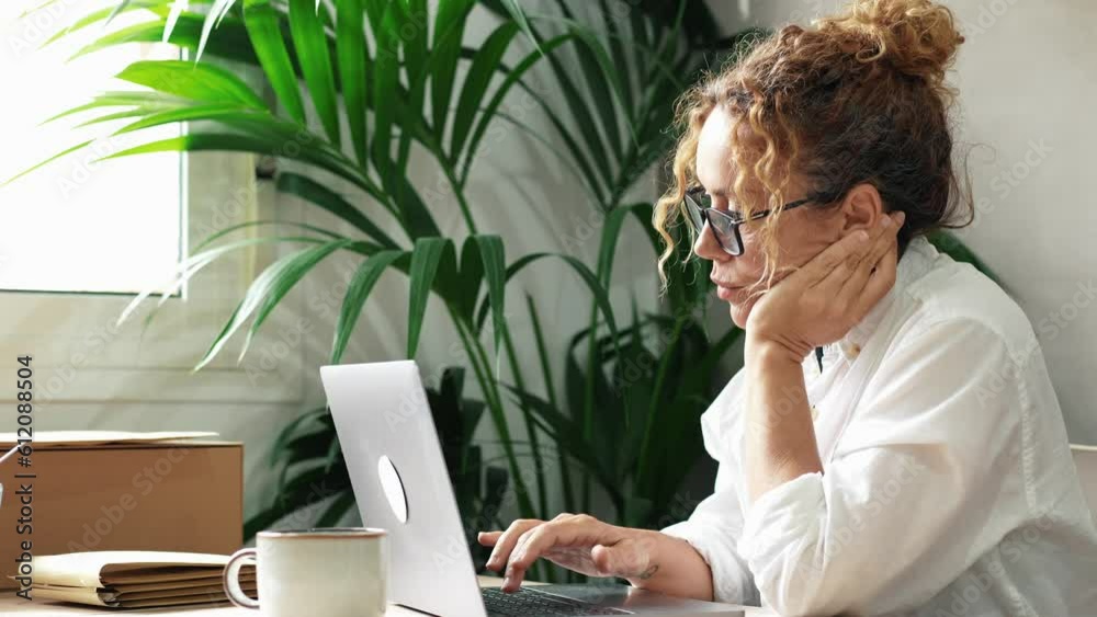 Modern woman entrepreneur doing boring work at laptop. Online job and boredom. People work in indoor home office workplace no happy. Tired and stressed lady using notebook on the table with plant back