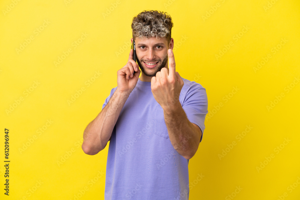 Young caucasian man using mobile phone isolated on yellow background doing coming gesture Young caucasian man using mobile phone isolated on yellow background doing coming gesture