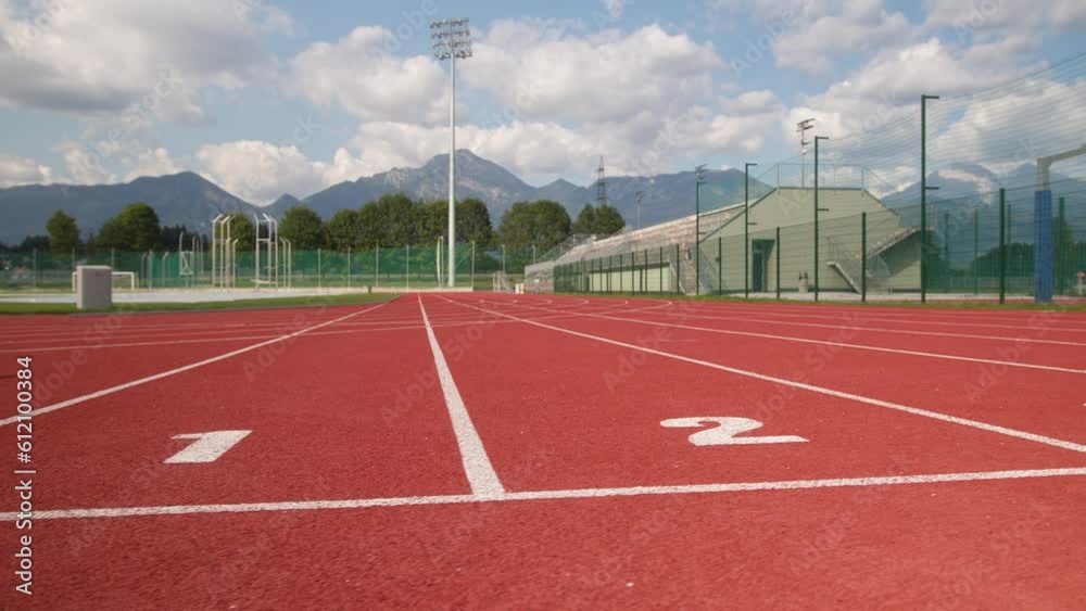 Empty red athletic track with seven-run lanes under a cloudy blue sky with mountains in the background, dolly right shot.
