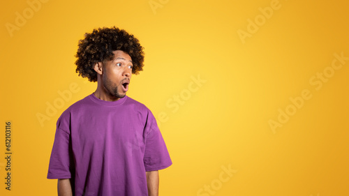 Cheerful shocked black adult curly man in purple t-shirt with open mouth looks at empty space