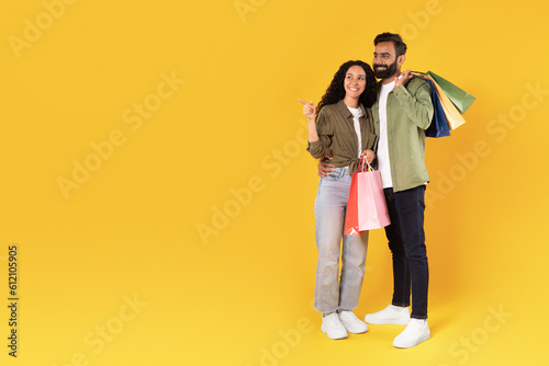 Middle Eastern Customers Couple With Bags Pointing Aside, Yellow Background