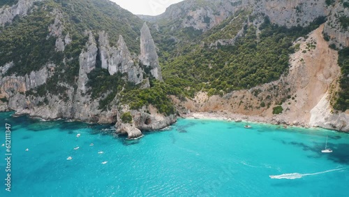 Aerial view of rocks and mountains, sea bay, beach, boats in summer day. Cala Goloritze, Sardinia Island, Italy. Top drone view of cliffs, stones, green trees, transparent azure water at sunset
