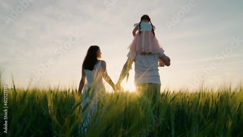 Young mother, father and little daughter play, enjoy nature outdoor, kids dream of flying. Mom, dad, girl child walk together, family of farmers with child on their shoulders walks through wheat field photo