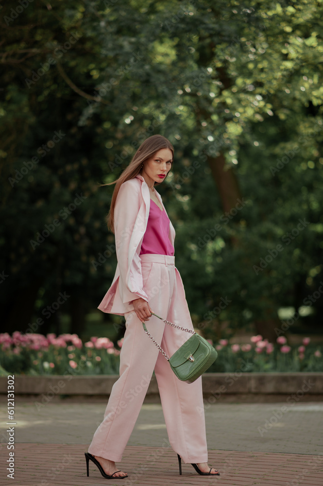 Fashionable elegant woman wearing trendy pink suit with classic blazer, wide leg trousers, high heeled sandals, holding green faux leather bag, walking in street. Full-length outdoor fashion portrait