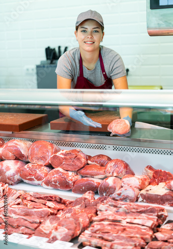 Smiling skilled young female butcher working behind counter in butchery, showing fresh raw veal loin slices