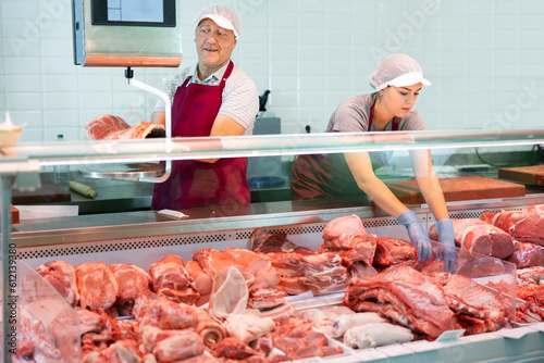 Experienced aged butchery shop owner working with young female assistant behind counter, weighing slab of meaty pork ribs on scales