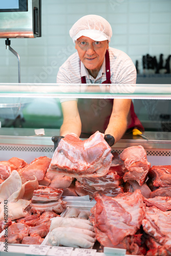 Positive skilled elderly butcher shop seller arranging meat products in glass refrigerated display case, laying out slab of fresh raw pork ribs