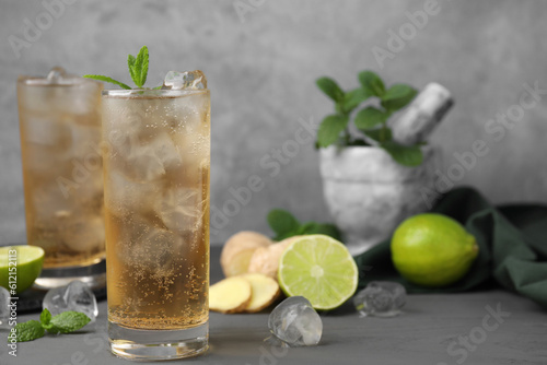 Glass of tasty ginger ale with ice cubes and ingredients on grey wooden table, space for text photo