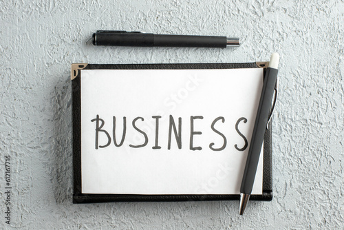 Top view of BUSINESS writing on white sheet and pen on new black closed notebook on gray sand background