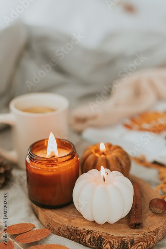Atmospheric candles - shape of pumpkins and candle in amber glass jar with wooden wick, autumn decor on gray bed. Autumn cozy home and hygge concept