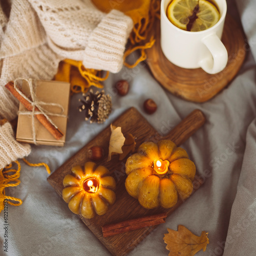 Top view on burning candles shape of pumpkins, mug with lemon tea, cozy warm sweater on bed. Autumn cozy home and hygge concept