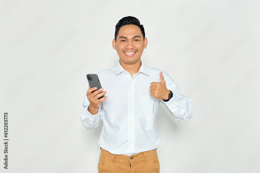 Portrait of smiling young Asian man in formal wear holding mobile phone and giving thumbs up, looking at camera with happy expression isolated on white background Portrait of smiling young Asian man in formal wear holding mobile phone and giving thumbs up, looking at camera with happy expression isolated on white background