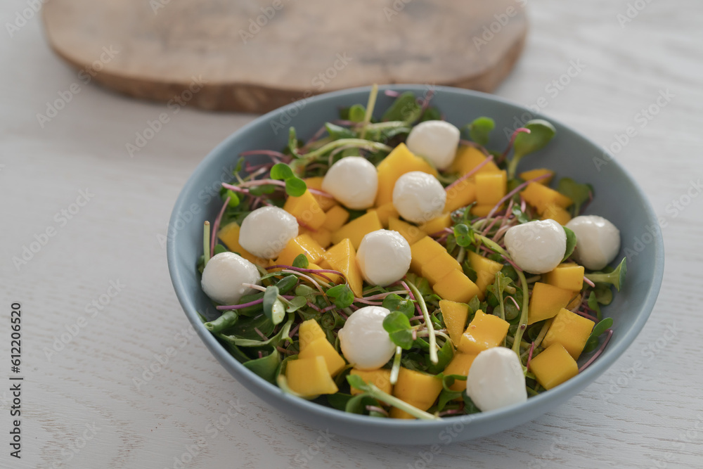 Salad with mango, mozzarella and mixed microgreens in blue bowl on white oak table Salad with mango, mozzarella and mixed microgreens in blue bowl on white oak table